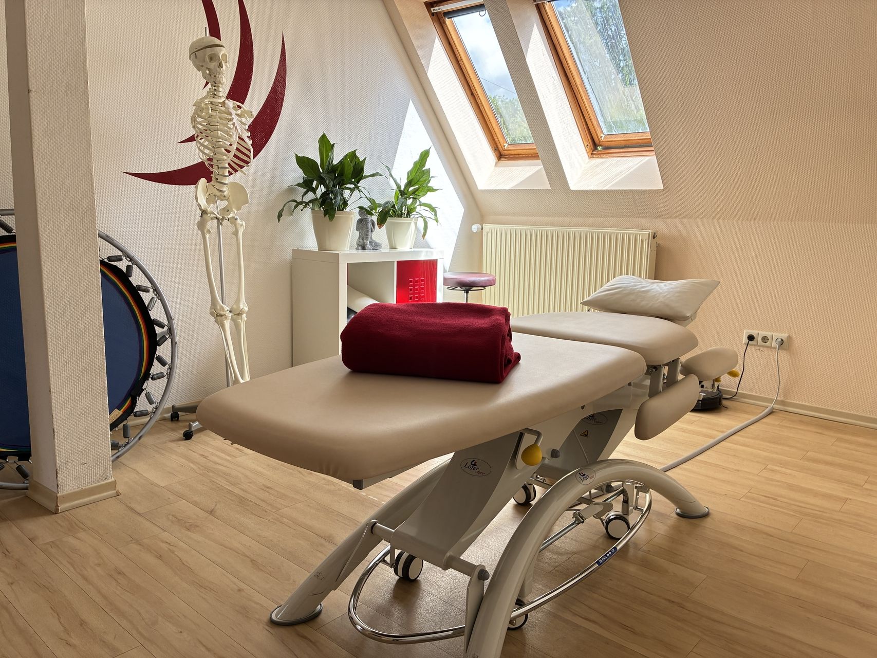 Behandlungsraum mit verstellbarer Liege, roter Decke, Skelettmodell, Pflanzen und Dachfenstern